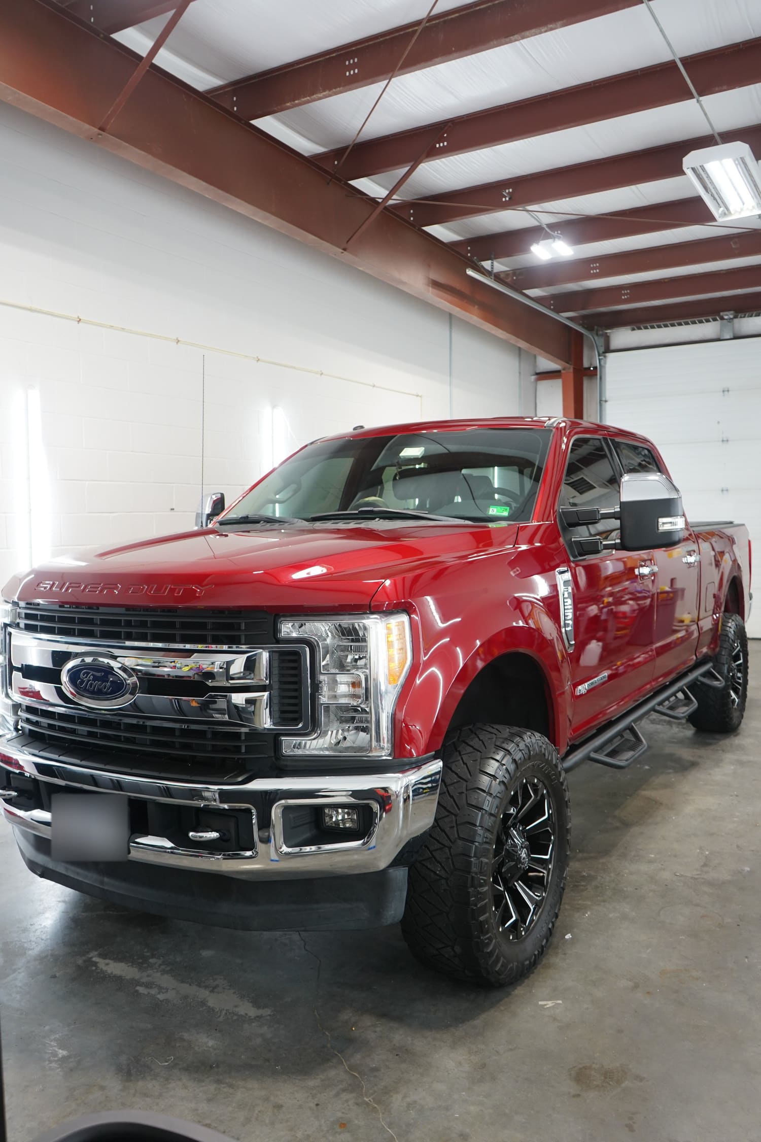 A red Ford Super Duty pickup truck parked inside a garage with a concrete floor, exposed metal beams on the ceiling, and a white wall with vertical lighting.