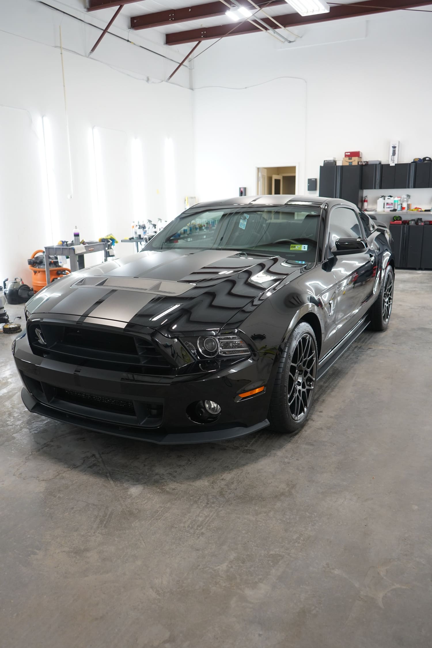 Black sports car, likely a Ford Mustang, parked inside a garage with tools and storage cabinets in the background.