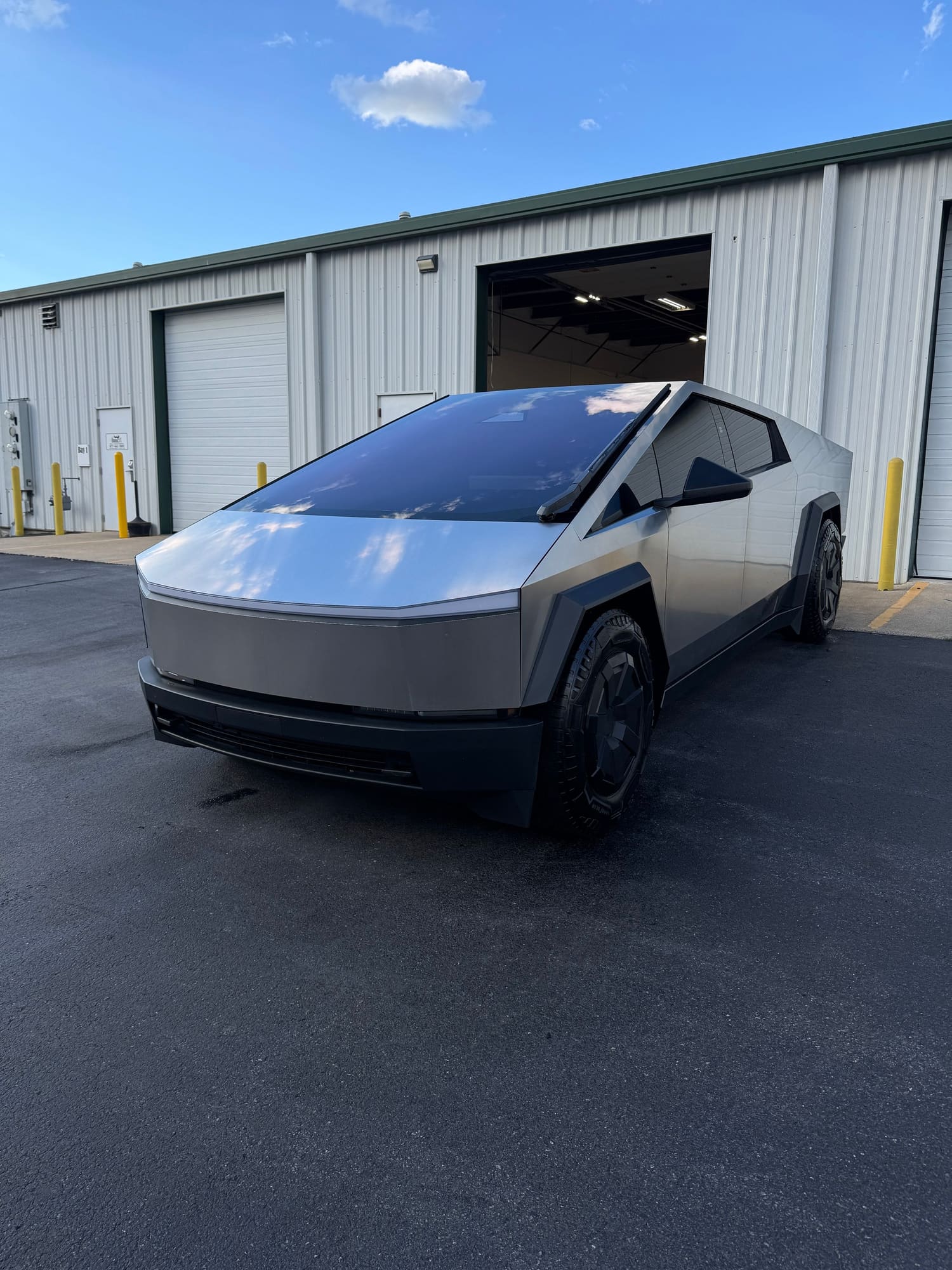 A futuristic silver vehicle with a sleek, angular design parked outside a warehouse with open garage doors under a blue sky with clouds.