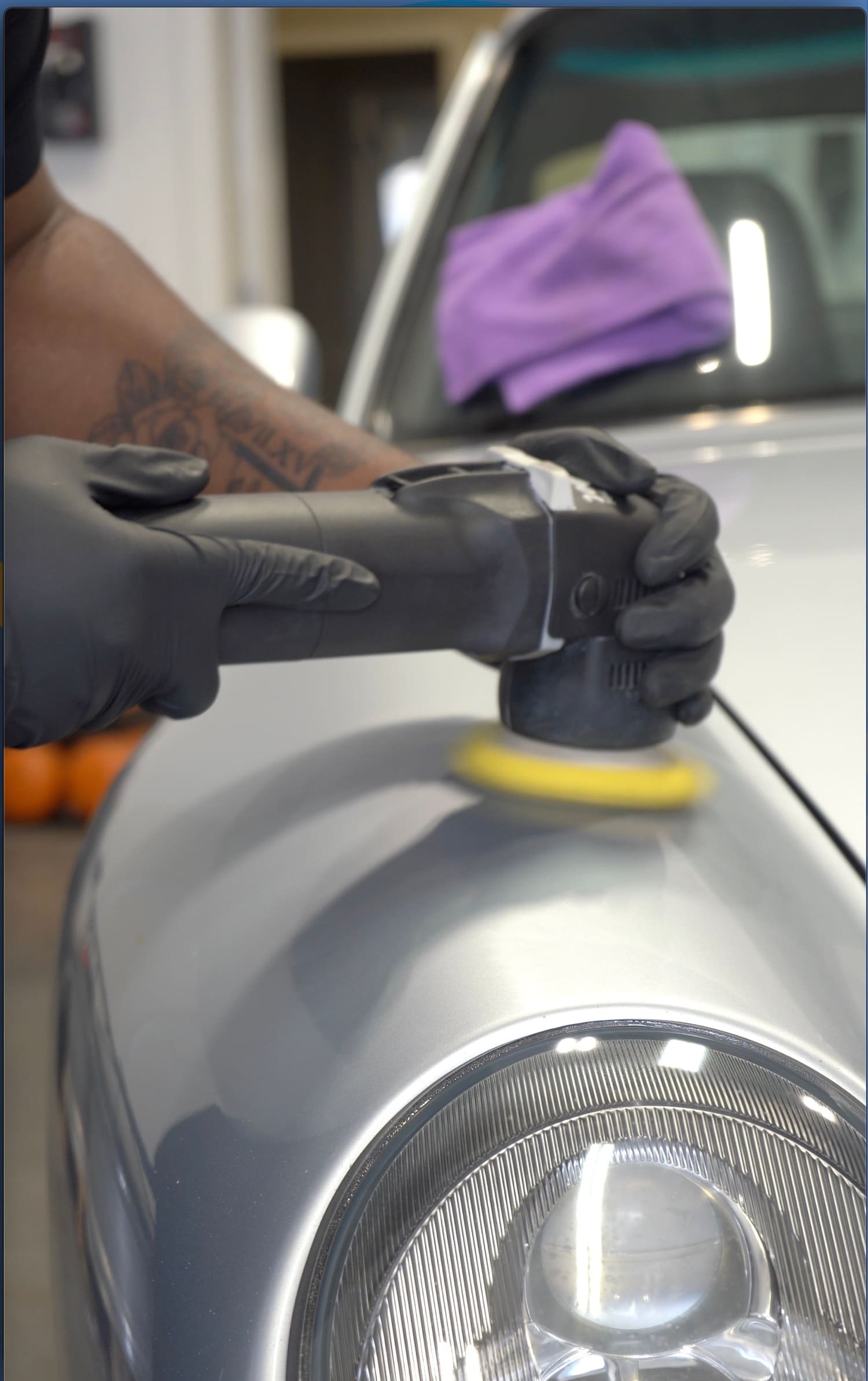 A person wearing black gloves using a polishing tool on the front fender of a silver car, with a purple cloth and headrest visible in the background.