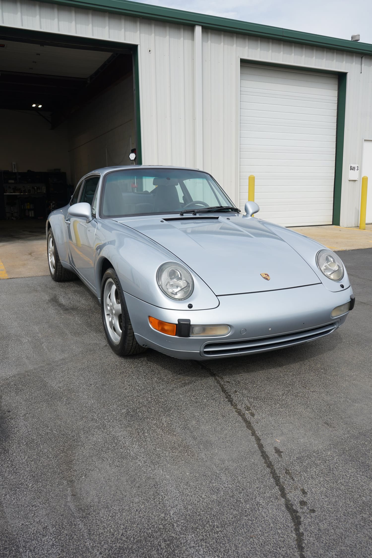 A silver Porsche 911 sports car parked outside a warehouse with a large open garage door and a white metal exterior.