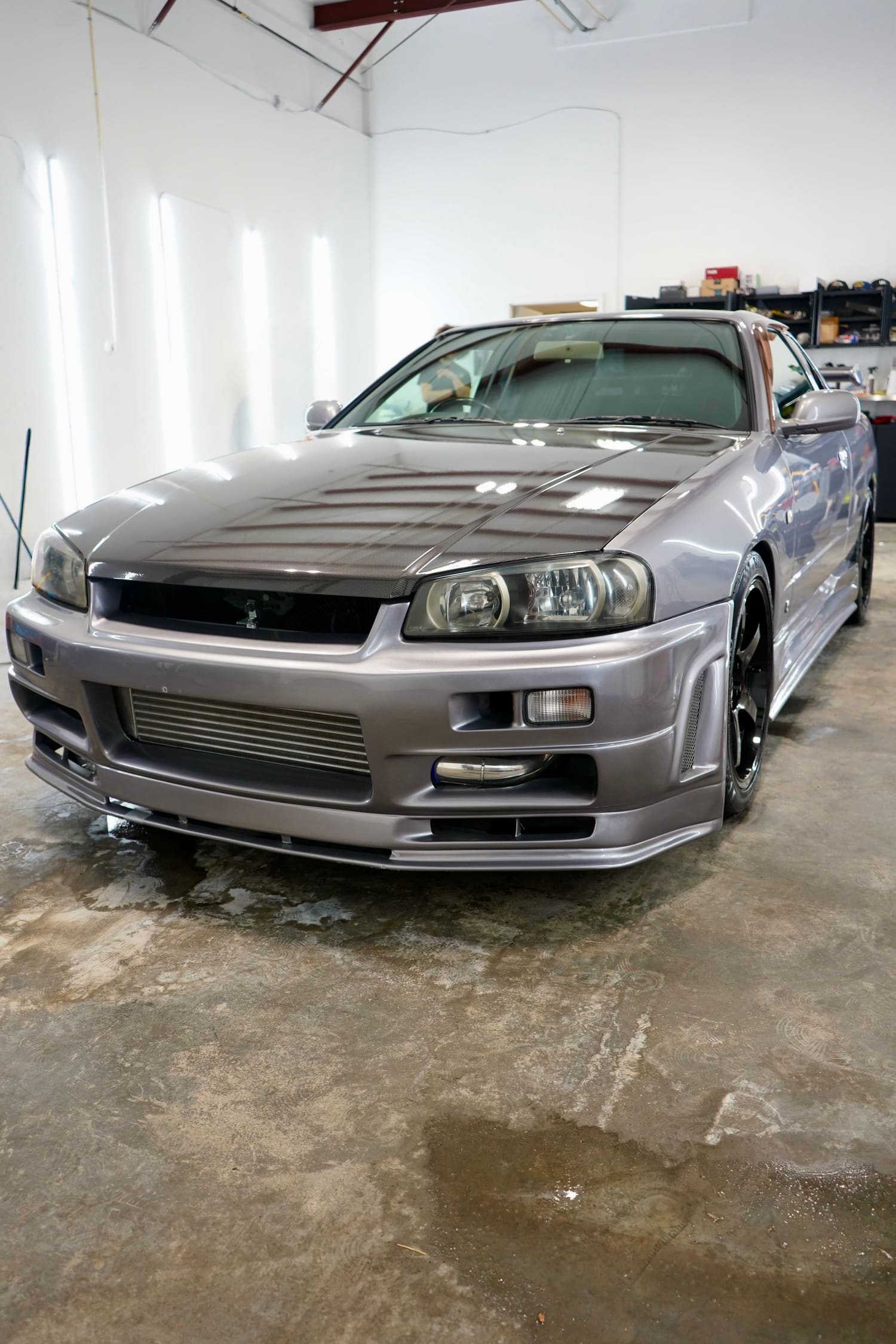 A silver sports car, likely a Nissan Skyline R32, is parked in a garage with a carbon fiber hood and aftermarket wheels.