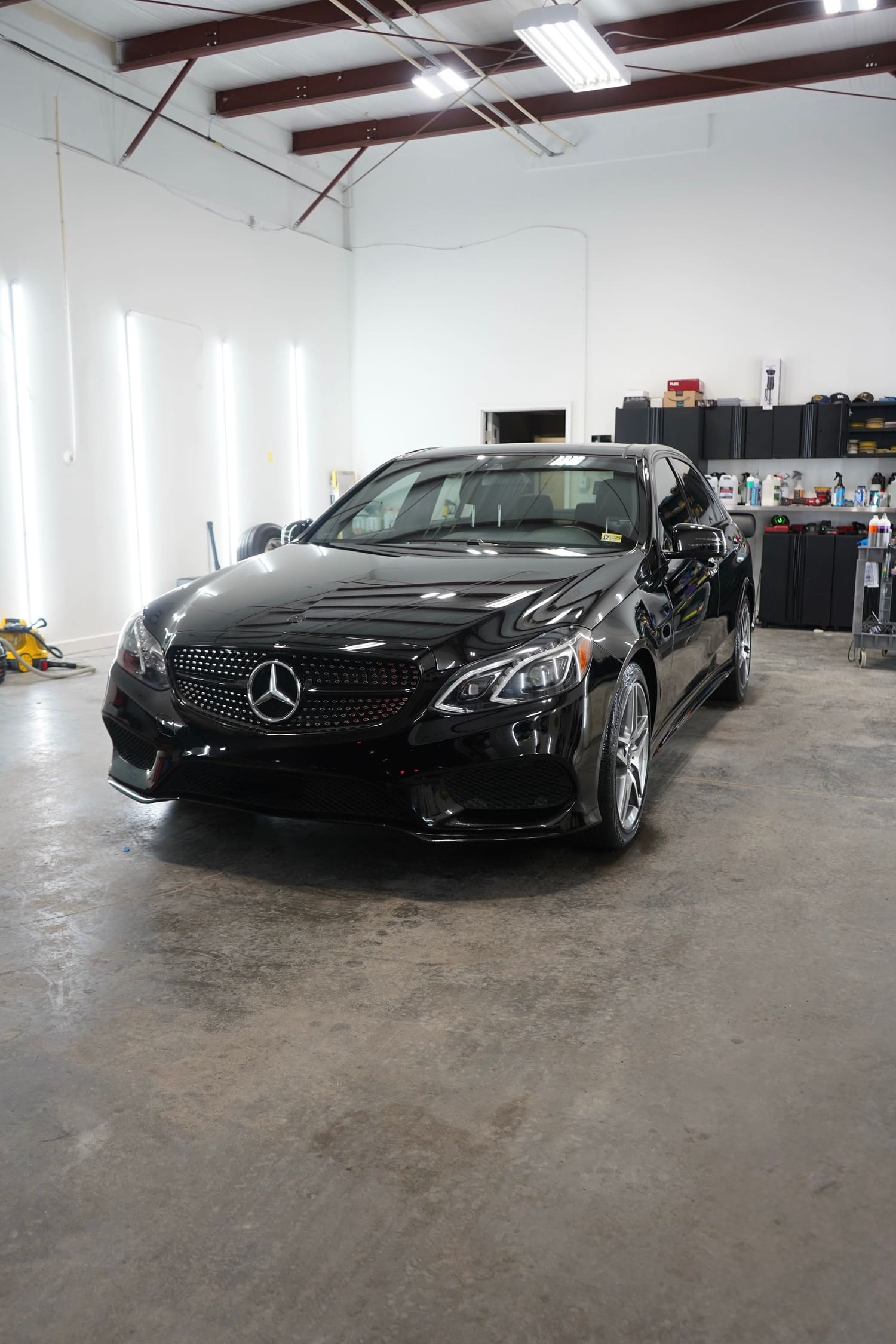A black Mercedes-Benz sedan parked inside a garage with white walls and a concrete floor. The garage has a high ceiling with exposed beams and fluorescent lighting. Shelves with various supplies are visible in the back right corner.