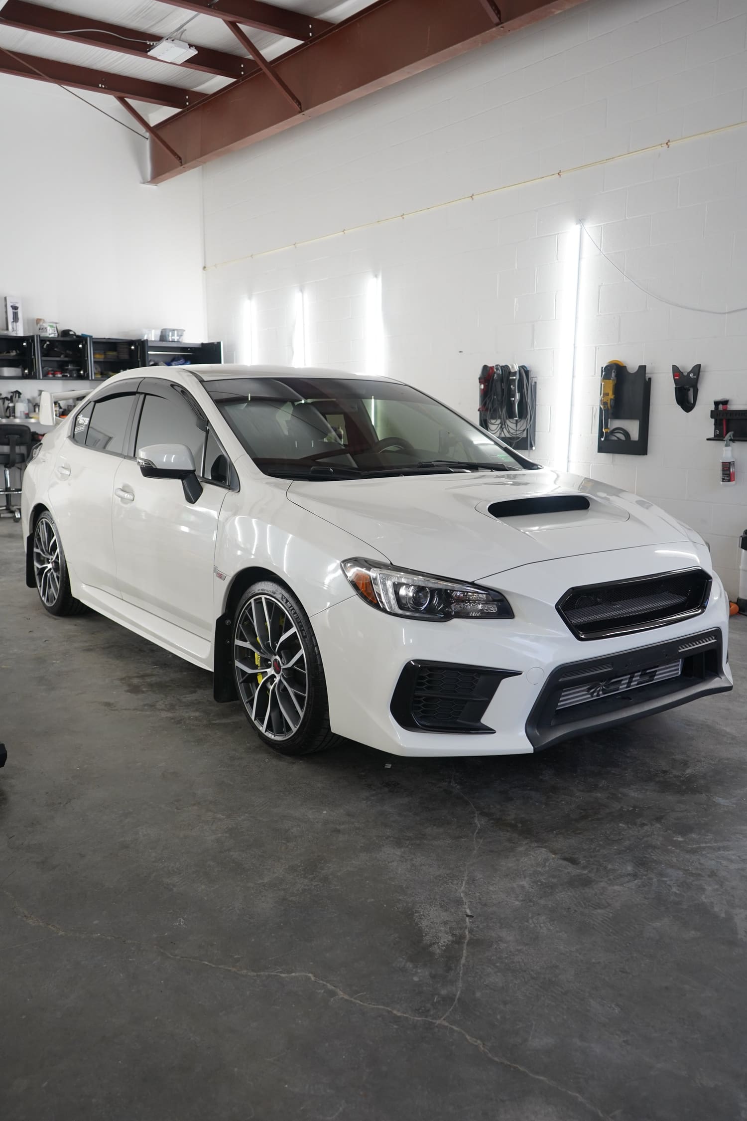 White Subaru WRX parked inside a garage with tools and shelves on the wall.