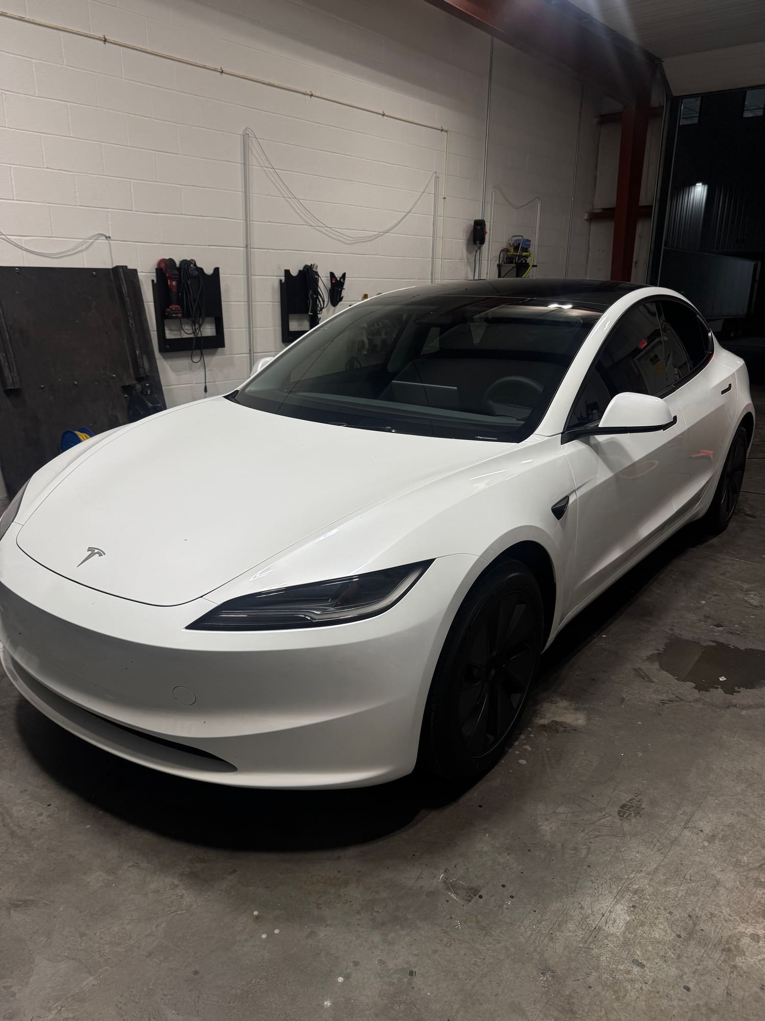 A white Tesla car parked inside a garage with tools and equipment mounted on the wall in the background.