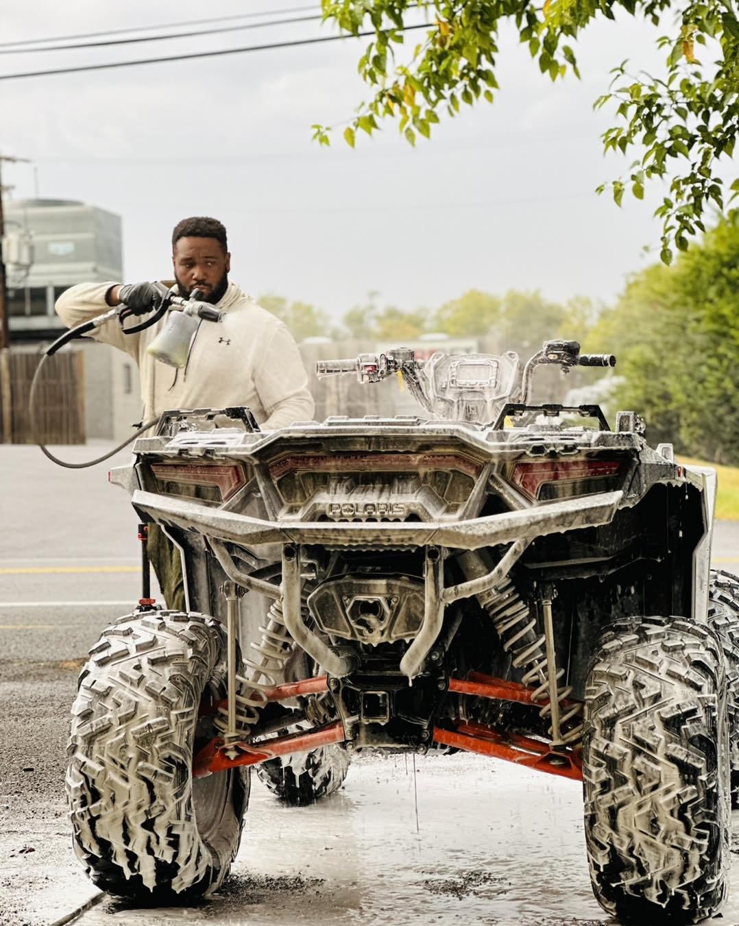 A man washing a muddy all-terrain vehicle on a road with greenery in the background.