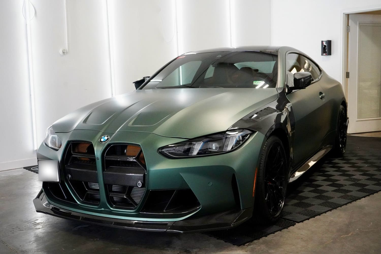 A green BMW sports car parked indoors on a black textured mat, with sleek aerodynamic design, black wheels, and a distinctive front grille.