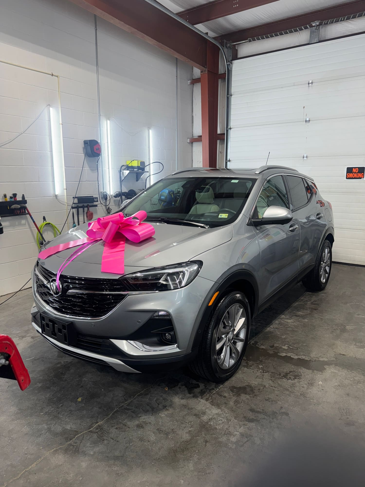 A silver SUV with a large pink ribbon and bow on the hood, inside a garage with white walls and a large rolling door.
